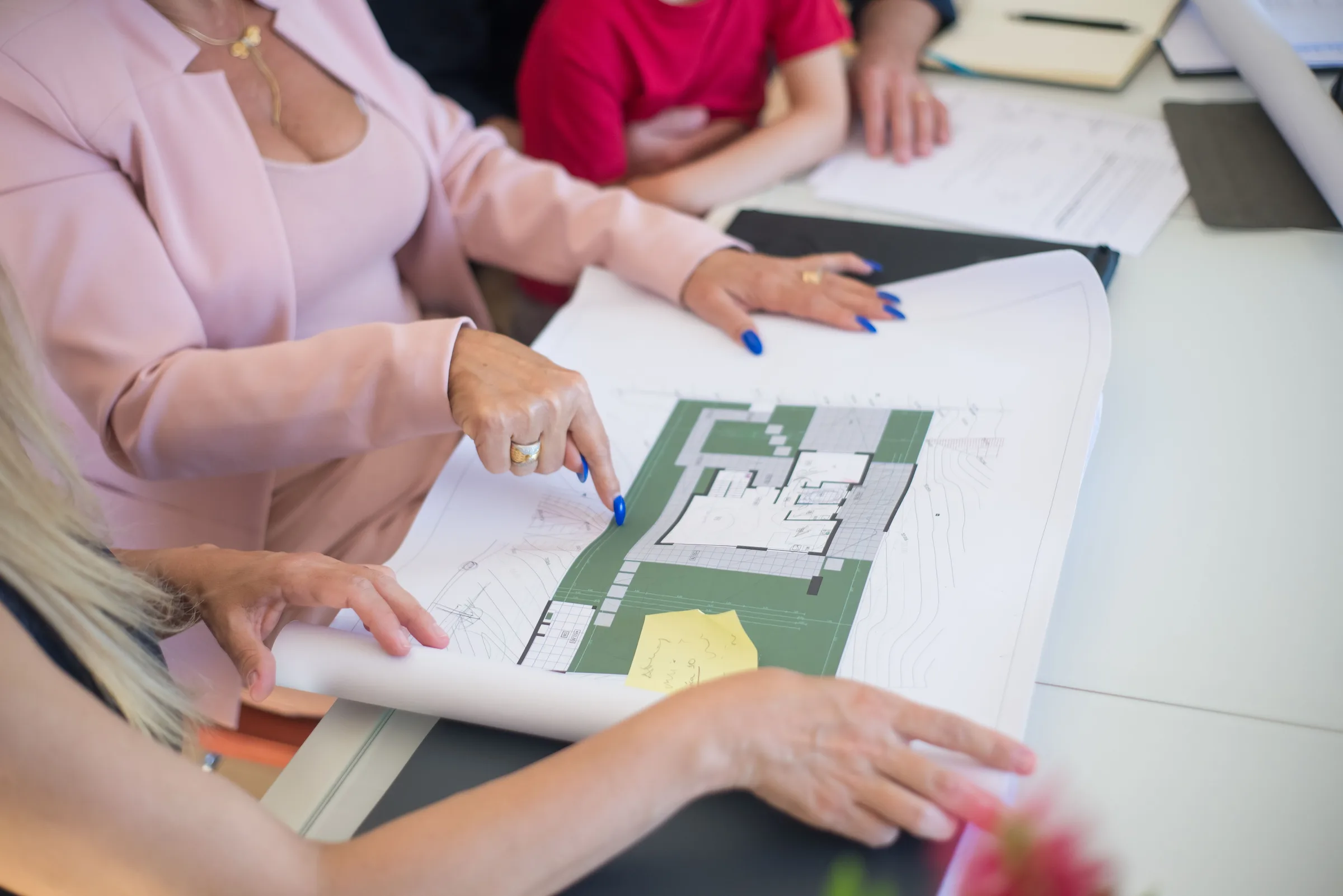 A real estate agent in a pink blazer shows floor plans to clients, discussing property details.
